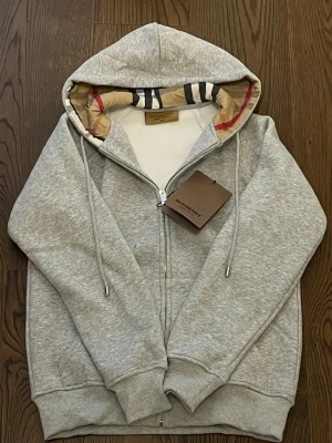 Grå zip hoodie från Burberry - Snygg grå hoodie från Burberry med dragkedja och klassiskt rutigt foder i huvan. Hoodien har långa ärmar, ribbade muddar och dragsko i huvan. Perfekt för dig som gillar stilrena plagg med exklusiva detaljer.