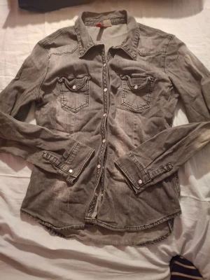 Grå jeansjacka från Divided H&M - Snygg grå jeansjacka från Divided by H&M med klassisk krage, två bröstfickor med lock och vita knappar. Figurnära modell med långa ärmar och dekorativa sömmar. Perfekt för en cool och avslappnad stil.