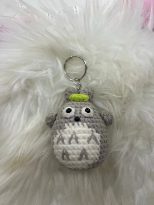 Virkat Totoro nyckelring grå - Söt handvirkad nyckelring i form av Totoro, gjord i grått och vitt garn med små detaljer i svart och grönt. Perfekt att hänga på väskan eller nycklarna för en gullig touch. Figuren har stora ögon och små öron, och är rund i formen.