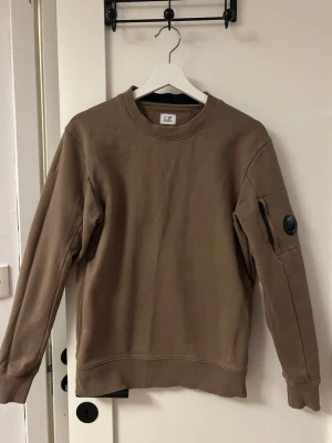 Brun/beige sweatshirt från C.P. Company - Säljer en brun/beige sweatshirt från C.P. Company med rund halsringning och ribbade muddar. Tröjan har en ikonisk svart linsdetalj på ärmen samt en liten ficka med dragkedja. Perfekt för dig som gillar streetwear och stilrena plagg.
