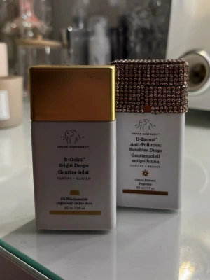 Drunk Elephant highlighter drops - Två highlighter drops från Drunk Elephant: B-Goldi Bright Drops med guldlock och D-Bronzi Anti-Pollution Sunshine Drops med ett lock dekorerat med glittrande stenar. Båda kommer i vita fyrkantiga flaskor, 30 ml, och ger glow och lyster till huden. Båda helt fulla med produkt. Endast testade 