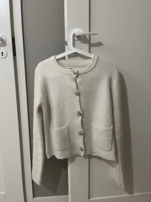 Vit kofta från Sinsay med fickor och guldknappar - Mysig vit kofta från Sinsay med rund halsringning och dekorativa guldfärgade knappar. Koftan har två praktiska fickor framtill och ribbade muddar vid ärmslut och nederkant. Perfekt att slänga på sig när det är lite kyligt ute. Passar som en s och xs också!