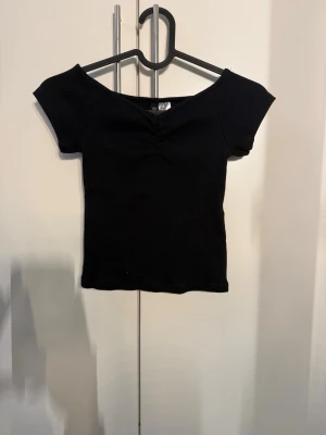 Svart ribbad offshoulder topp - Snygg svart ribbad topp med offshoulder-design och korta ärmar. Toppen har en lätt v-ringad form framtill och sitter tight mot kroppen för en stilren look. Perfekt att matcha med jeans eller kjol för en trendig outfit.