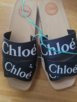 Svarta sandaler från Chloé - Snygga svarta sandaler från Chloé med två breda band i textil med vit logotyp. Sandalerna har fyrkantig tå och platåsula i flätad jute för en trendig espadrille-look. Perfekta för sommaren och ger en cool touch till din outfit.