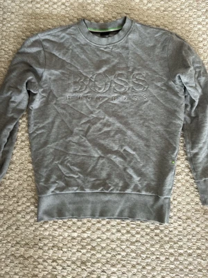 Grå sweatshirt från Hugo Boss - Snygg grå sweatshirt från Hugo Boss med broderad logga på bröstet. Tröjan har rund halsringning, ribbade muddar vid ärmslut och nederkant samt en klassisk passform. Perfekt för en avslappnad och stilren look.