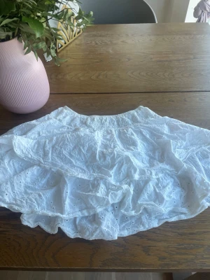 Vit kort kjol med volang och broderi - Supersöt vit kort kjol med volang och broderade detaljer. Kjolen har resår i midjan för skön passform och är gjord i ett lätt bomullstyg, perfekt för varma dagar. Broderade hålmönster ger en romantisk och luftig känsla.