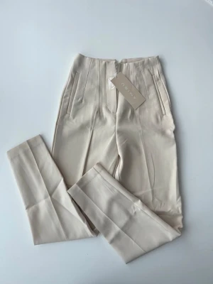 Beige kostymbyxor från Amisu - Stilrena beige kostymbyxor från Amisu med raka ben och pressveck. Byxorna har hög midja, två sidofickor och stängs med dragkedja och hake. Materialet är mjukt och har en lätt glansig finish som ger en snygg look.