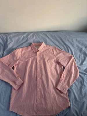 Röd/rosa vit randig skjorta med knappar - Säljer en klassisk röd/vit randig skjorta med långa ärmar och knappar framtill. Skjortan har en snygg krage och smala ränder som ger en fräsch look. Perfekt till jeans eller chinos för en clean stil.