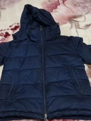 Mörkblå pufferjacka med huva - Snygg mörkblå pufferjacka med dragkedja framtill och två stora fickor med dragkedja. Jackan har en hög krage och en rejäl huva som skyddar mot kyla. Perfekt för kalla vinterdagar och ger en sportig look.