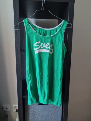 Grön linnetopp med vit text - Snygg grön linnetopp med vit text 'Svez' framtill och vita kantband. Perfekt för varma dagar och enkel att matcha med jeans eller shorts. Luftig och bekväm passform.