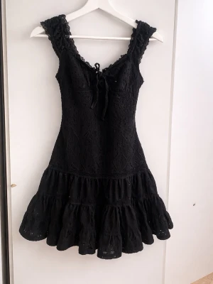 Svart spetsklänning med snörning🖤 - Svart kortklänning med spetsdetaljer och snörning framtill. Klänningen har volang nertill och är ärmlös med v-ringad hals. Perfekt för dig som vill ha en feminin och trendig look.