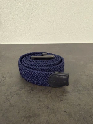 Marinblått flätat bälte från Gant, stl 115cm - Snyggt marinblått flätat bälte från Gant med detaljer i svart skinn. Bältet har en klassisk metallspänne och är perfekt för att lyfta vilken outfit som helst. Märket syns diskret på skinnpartiet vid änden.
