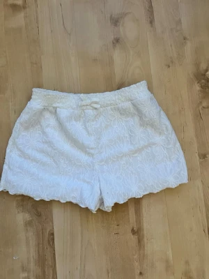 Vita spetsmönstrade shorts med snörning - Säljer ett par vita shorts med spetsmönster och elastisk midja. Shortsen har en liten snörning framtill och vågig kant nedtill för en extra detalj. Perfekta för varma dagar och ger en fräsch och somrig look.