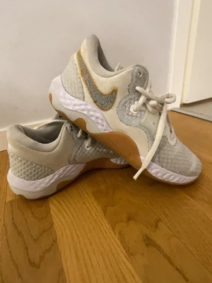 Nike vita sneakers med beige detaljer - Snygga vita sneakers från Nike med beige och grå detaljer. Skorna har ett stickat meshmaterial för bra andningsförmåga, vit sula med gummidetaljer och klassisk Nike Swoosh i silver och guld. Perfekta för en sportig och trendig look.