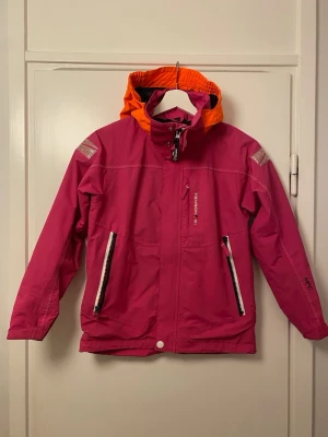 Rosa vindjacka från Tenson - Snygg rosa vindjacka från Tenson med orange huva och vita dragkedjor. Jackan har två framfickor med dragkedja, reflexdetaljer på axlarna och justerbar huva. Perfekt för blåsiga dagar och ger en färgklick till din outfit.