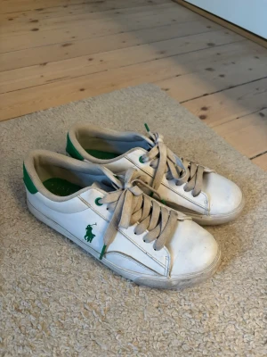 Polo Ralph Lauren sneaker - Snygga vita sneakers från Polo Ralph Lauren med gröna detaljer och logga på sidan. Klassisk låg modell med vita snören och gummisula. Perfekta för en avslappnad och sportig stil. Tillverkade i läder med textilfoder.