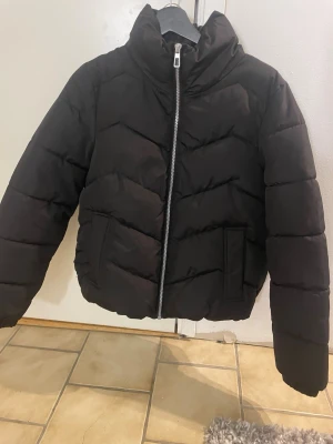 Svart pufferjacka från Vero Moda - Snygg svart pufferjacka från Vero Moda med hög krage och silvrig dragkedja. Jackan har två stora fickor framtill och quiltad design i breda paneler. Perfekt för kalla dagar och ger en trendig look. Materialet är syntetiskt och jackan är kort i modellen.