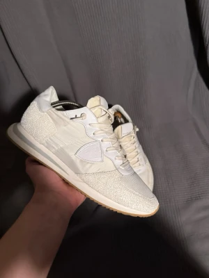  Philippe Model - Snygga vita sneakers från Philippe Model med glittriga detaljer och klassisk logotyp på sidan. Skorna har en mix av textil och mocka, vita skosnören och en bekväm sula i gummi. Perfekta för dig som vill ha en clean och trendig look.