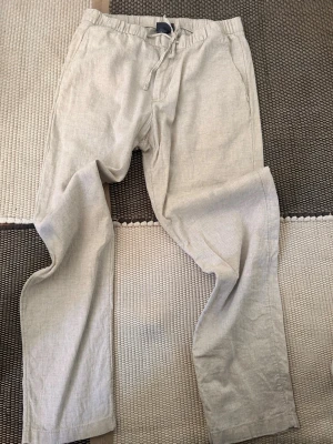 Beiga linnebyxor med dragsko - Säljer ett par beiga linnebyxor med avslappnad passform och dragsko i midjan. Byxorna har raka ben och sidofickor, perfekta för en chill och stilren look. Materialet är luftigt linne som känns svalt och skönt.