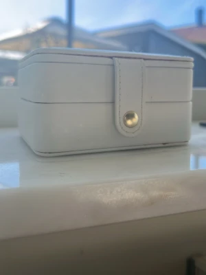 Vit smyckesbox med spegel - Snygg vit smyckesbox i fyrkantig form med guldfärgad knappstängning. Insidan har flera fack för ringar och småsaker samt en spegel i locket. Perfekt för att organisera smycken eller accessoarer. Tillverkad i konstläder.