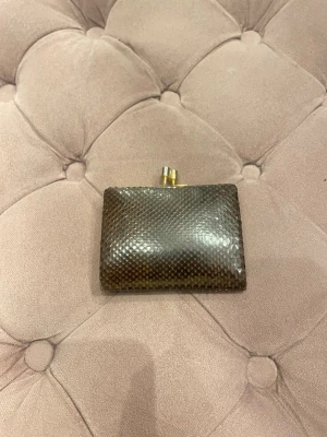 Brun plånbok i ormmönstrat läder - Säljer en brun plånbok i ormmönstrat läder med guldfärgad metallram och klassiskt trycklås. Plånboken har en stilren och kompakt design som gör den enkel att ta med sig. Perfekt för dig som gillar en unik accessoar med vintagekänsla.