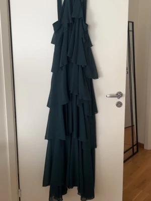 Mörkgrön långklänning med volanger - Säljer en elegant mörkgrön långklänning med flera lager av volanger och en dekorativ rosdetalj vid axelbanden. Klänningen har ärmlös design och är tillverkad i ett lätt och flödande syntetmaterial som ger ett snyggt fall.