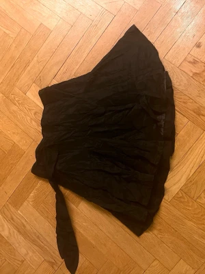 Svart omlottkjol med volang - Säljer en svart kort omlottkjol med volangkant. Kjolen har en knytning i sidan och är gjord i ett lätt, något glansigt tyg som ger en snygg struktur. Perfekt för dig som gillar trendiga och feminina detaljer.Skriv gärna för mått! 💕