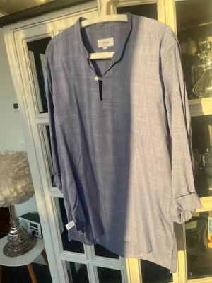 Blå henley-skjorta från Eton - Säljer en stilren blå tun sommar skjorta från Eton med diskret rutigt mönster. Skjortan har lång ärm, knappar vid halsen och är tillverkad i ett lätt och luftigt tyg, perfekt för en avslappnad men snygg look. Modellen är utan manschetter, utan viks upp snyggt