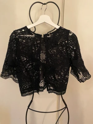 Svart spetskofta med kort ärm - Snygg svart kofta i spets med korta ärmar och rund hals. Koftan är använd fåtal gånger och inga slitage. Knäpps med en knapp fram. Perfekt att slänga över ett linne eller klänning. 