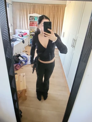 Svart omlottopp med långa ärmar - Snygg svart omlottopp med långa ärmar och knytband i midjan. Tröjan har djup v-ringning och ribbad struktur som ger en trendig look. Perfekt att styla med höga jeans eller kjol för en cool outfit.