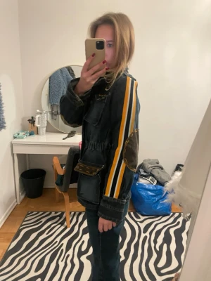 Mörk jeansjacka med patchar och ränder - Unik mörkblå jeansjacka med coola patchar, bland annat dödskalle och text på bröstet. Jackan har gula och vita ränder längs ärmarna, bruna detaljer på armbågar och fickor samt flera stora fickor framtill. Snygg streetstil och perfekt för dig som vill sticka ut.