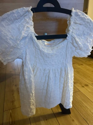 Vit topp med puffärmar - Supersöt vit topp med puffiga, korta ärmar och strukturerad, rynkad textur. Toppen har en rund halsringning och en luftig passform som ger en trendig och ungdomlig look. Perfekt för sommarens alla tillfällen!