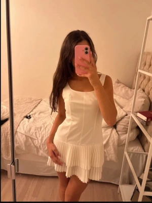 Vit kort klänning med plisserad kjol - Säljer en söt vit kort klänning med breda axelband och plisserad kjol. Klänningen har en enkel, rak överdel och två lager volang nedtill som ger extra volym. Den har inbyggda shorts! Perfekt till studenten 