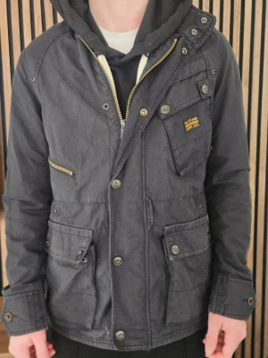 Svart parkajacka från G-Star Raw - Cool svart parkajacka från G-Star Raw med flera fickor, knappar och dragkedjor. Jackan har en hög krage, långärmad design och detaljer som logopatch på bröstet. Perfekt för dig som vill ha en stilren och funktionell jacka.