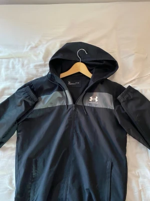 Svart vindjacka från Under Armour - Snygg svart vindjacka från Under Armour med huva och dragkedja framtill. Jackan har en grå panel över bröstet och vitt logotryck på vänster sida. Tillverkad i lätt, vindtätt material med meshfoder för extra komfort.