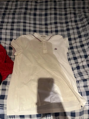 Vit Moncler pikétopp med ränder - Klassisk vit pikétopp från Moncler med korta ärmar och knappar framtill. Kragen har tunna röda och blå ränder och Moncler-logga på bröstet. Perfekt till jeans eller kjol för en clean och sportig look. Obs äkta