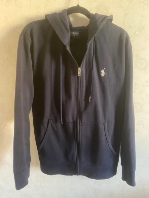 Navy Ralph Lauren Zip-Up - Säljer en mörkblå hoodie från Polo Ralph Lauren med dragkedja och två fickor framtill. Klassisk logga med broderad ryttare på bröstet. Hoodien har huva med snören och är tillverkad i mjukt bomullsmaterial. Perfekt för dig som gillar stilrena och bekväma plagg.