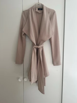 Beige trenchcoat från ONLY - Stilren beige trenchcoat från ONLY med långa ärmar, bred krage och knytskärp i midjan. Jackan har en mjuk, slät yta och ett elegant fall. Perfekt för dig som gillar minimalistisk och modern stil.