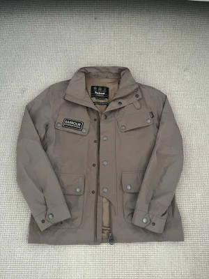 Barbour International jacka – nyskick - Barbour International jacka – nyskick  Säljer en stilren och funktionell jacka från Barbour International i en snygg beige/taupe färg. Jackan är endast använd 2–3 gånger och är i princip i nyskick – inga fläckar, slitage eller defekter.  Perfekt som vår- och höstjacka med flera praktiska fickor och en tidlös design som passar både till vardag och lite mer uppklätt.  Detaljer: 	•	Storlek: (fyll i din storlek här) 	•	Skick: Nästan ny 	•	Färg: Beige/Taupe 	•	Originalkvalitet från BarbourInternation
