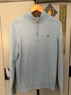 Ljusblå halvzip-tröja Morris - Ljusblå stickad tröja från Morris med halv dragkedja och ståkrage. Tröjan har ett diskret Morris-emblem broderat på bröstet och ribbade muddar vid ärmslut och nederkant. Perfekt för en stilren och avslappnad look. L men passar M