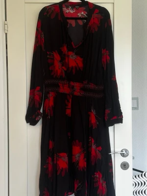 Svart långklänning med röda blommor Desigual - Långklänning från Desigual i svart med stora röda blommor. Klänningen har lång ärm, v-ringning och markerad midja med smockdetalj. Luftigt material och flowy passform, perfekt för dig som gillar färgstarka mönster.