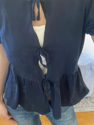 Mörkblå blus med knyt framtill - Snygg mörkblå blus med korta ärmar och tre dekorativa knytningar framtill. Blusen har en lätt rynkad midja som ger en avslappnad och trendig siluett. Tillverkad i ett mjukt tyg som känns skönt mot huden. Perfekt att matcha med jeans eller kjol.