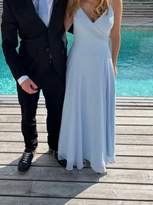 Ljusblå långklänning med snörning - Elegant ljusblå långklänning med smala axelband och v-ringning. Klänningen har en öppen rygg med snygg snörning och ett svepande, luftigt fall. Perfekt för student eller bal. Tillverkad i ett mjukt, tunt material som ger en fin siluett.