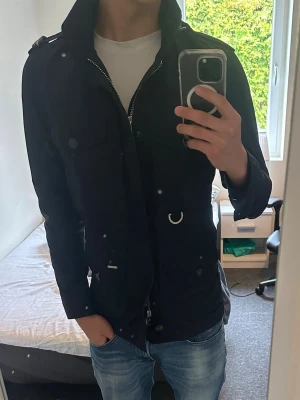 Field jacka från Snoot - Snygg marinblå jacka från Snoot med dragkedja och knappar framtill. Jackan har flera fickor, hög krage och detaljer i metall. Tillverkad i ett lätt, vattenavvisande material som passar perfekt till vår och höst.