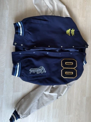 Blå och beige bomberjacka med tryck - Snygg bomberjacka i college-stil med marinblå kropp och beige ärmar. Jackan har broderade detaljer som siffran 80 på framsidan, 13 och X på ärmarna samt texten 'Official'. Ribbstickade muddar med blå och vita ränder. Perfekt för en sportig och trendig look. Köpt från manavdelningen men funkar även bra för tjejer