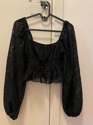Svart croppad spetsblus - Säljer en svart croppad blus med långa puffärmar och vacker spetsdetalj. Toppen har resår i midjan och vid ärmsluten för en snygg passform. Perfekt för dig som gillar romantiska och trendiga plagg.