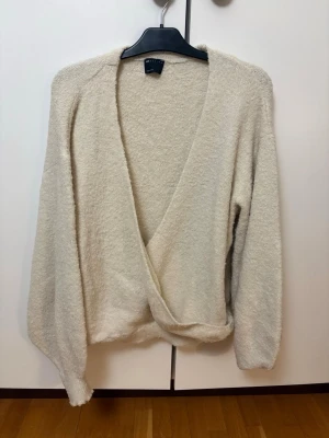 Beige omlottkofta från ASOS DESIGN - Mysig cremefärgad omlottkofta från ASOS DESIGN med lång ärm och djup v-ringning. Koftan har en loose passform och är tillverkad i ett mjukt, stickat material som känns skönt mot huden. Använd en gång. 