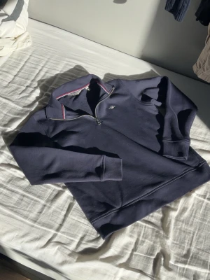 Mörkblå quarter zip från Gant - Snygg mörkblå quarter zip från Gant med liten logga på bröstet. Tröjan har hög krage med dragkedja och diskreta röda och vita detaljer på insidan av kragen. Tillverkad i mjukt bomullsmaterial och har långa ärmar.