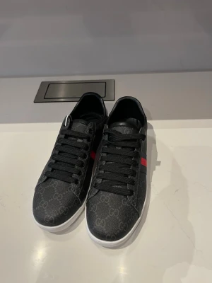 Svarta Gucci sneakers med röd/blå rand - Snygga svarta sneakers från Gucci med klassiskt GG-mönster över hela skon. Skorna har svarta skosnören, vit sula och en ikonisk röd och blå rand på sidan. Materialet är textil med läderdetaljer och insidan är svart. Perfekta för dig som gillar exklusiv streetstyle.
