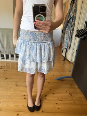 Blommig volangkjol i blått och vitt🫶🏻 - Söt kort kjol med blått och vitt blommönster och volangdetaljer. Kjolen har bred smock i midjan för skön passform och är gjord i ett lätt, luftigt material. Perfekt för varma dagar och enkel att matcha med olika toppar. Super söt!🤍🤍